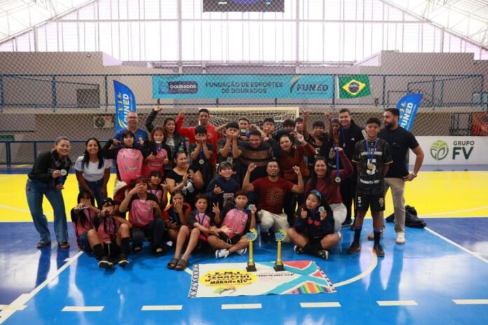 Group of children and adults posing with medals and two trophies after a sports event on a blue and yellow indoor court; banners and netting visible in the background.