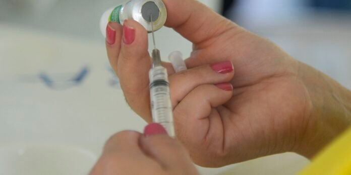 Close-up of hands drawing liquid from a vial into a syringe on a clinical surface, with pink nails visible.