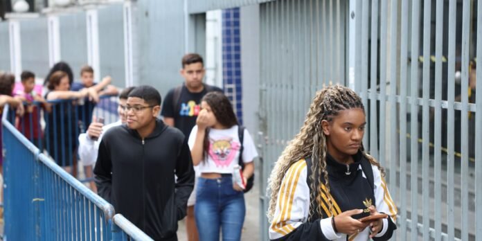 Young woman with braided hair in a black and yellow Adidas jacket checks her phone while walking past a blue railing; other students line up in the background.