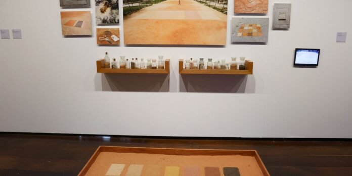 Gallery wall with a photo collage above two wooden shelves holding glass bottles; a sand tray with color samples sits on the floor in the foreground, and a small touchscreen panel is on the right.