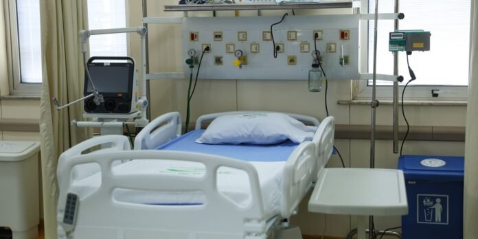 Hospital patient bed in a clinical room with wall-mounted medical outlets and monitoring equipment in the background, ready for use