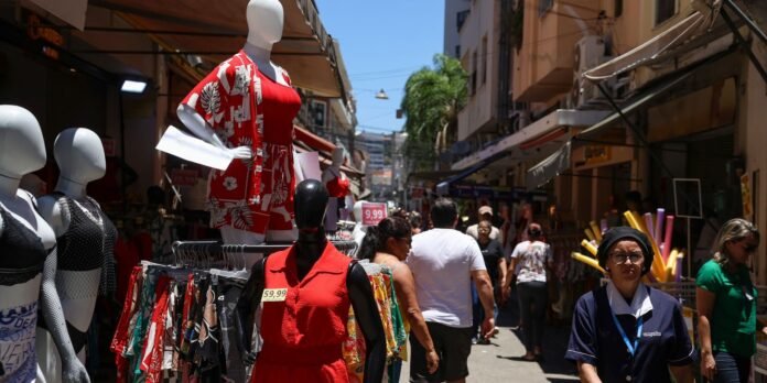Busy outdoor market with mannequins displaying red outfits among clothing racks and shoppers walking down a sunlit street