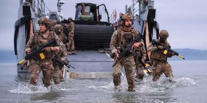 Soldiers in camouflage wading through shallow water as they disembark from a landing craft with rifles ready, during an amphibious operation.