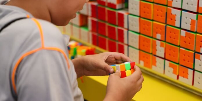 Person holding and twisting a colorful Rubik's cube in front of shelves filled with stacked puzzle boxes.