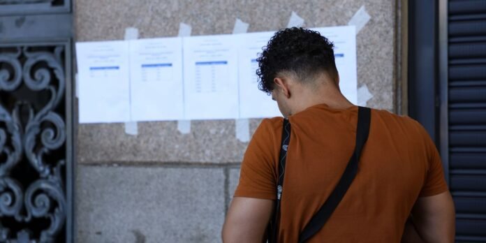 Man in an orange shirt with a shoulder strap reads posted notices taped to a stone-wall surface.