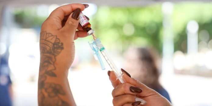 Close-up of a tattooed arm drawing vaccine liquid from a vial into a syringe with blue protective cap visible on the syringe clip.