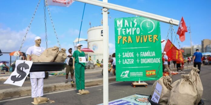 Street protest scene with a large green banner reading 'Mais Tempo pra Viver' and lists 'saúde, família, lazer, dignidade' in white letters; a hanging scale with burlap bags in the foreground and people in costumes in the background.
