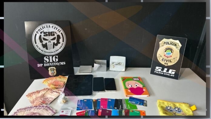 Seized items on a table: cash, several mobile phones, bank cards, a small pouch, a colorful notebook, and police signs SIG DP BONITO/MS and Polícia Civil-MS