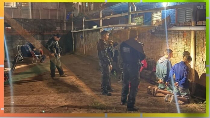 Police officers detain and surround several kneeling individuals against a brick wall in a dim alley at night, with a chair and bystanders in the background.