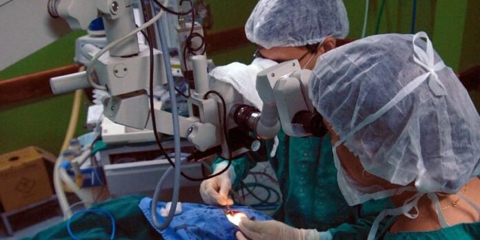 Surgeons in gowns and caps perform a delicate operation under a stereomicroscope in a sterile operating room area with drapes and equipment nearby.