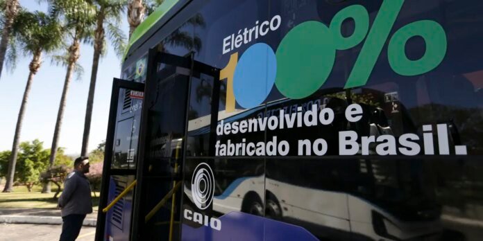 Blue promotional bus with large green percentage graphic and the words 'Elétrico' and 'desenvolvido e fabricado no Brasil' visible; man stands near an open bus door as palm trees line the background (CAIO logo also visible).