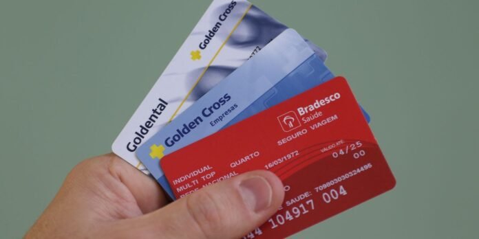 Hand holding several fanned-out health insurance cards (Golden Cross and Bradesco Saude) in red, blue, and white, against a green background.