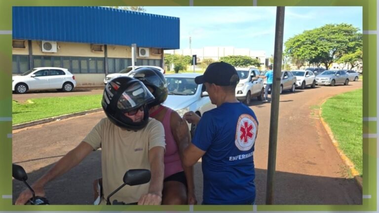 Medical worker in a blue shirt vaccinates a motorcyclist wearing a helmet at a drive‑through clinic, with cars in line behind them.