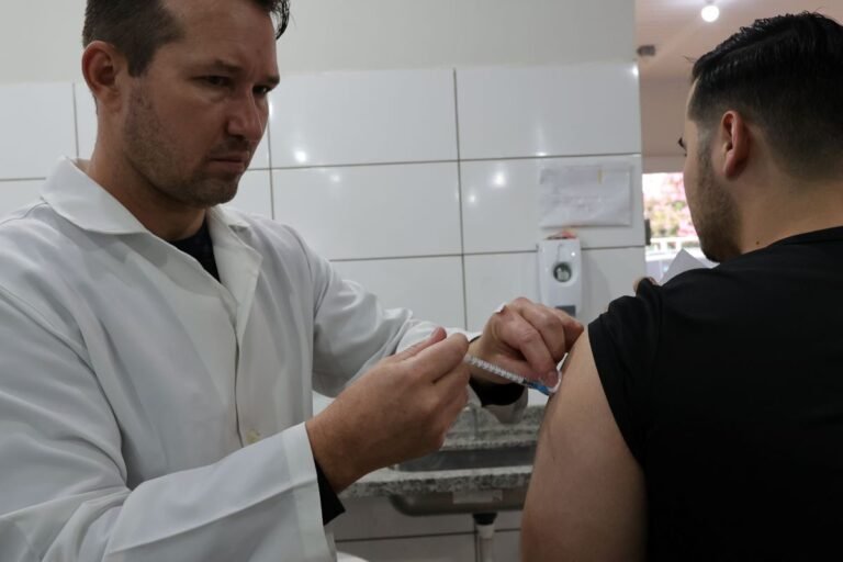 A clinician in a white coat administers a vaccination to a patient's upper arm with a syringe in a clinical setting