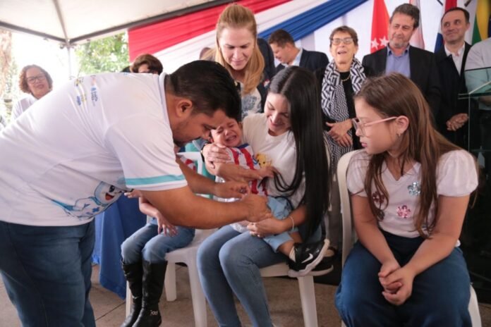 Man in a white shirt leans in to interact with a crying baby held by a smiling mother, as a teenage girl sits nearby and onlookers watch under a colorful canopy.