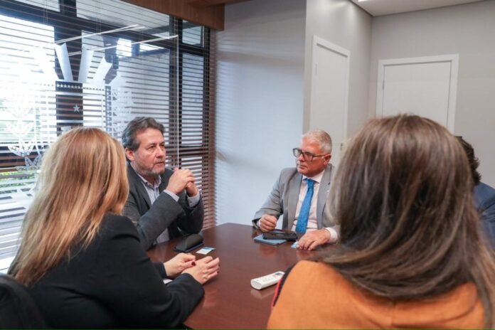 Group of professionals in a business meeting around a wooden conference table, one man speaking with hands clasped.