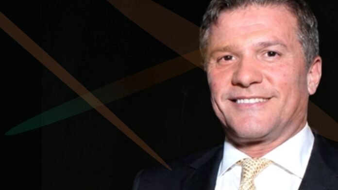 Smiling man in a dark suit and cream tie against a dark backdrop with diagonal overlay graphics.