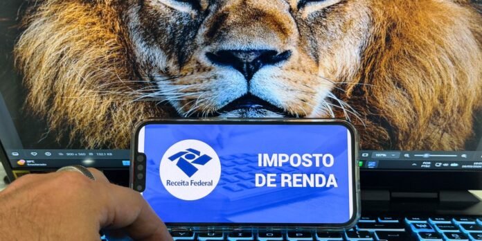 Smartphone screen displays 'Imposto de Renda' and the Receita Federal logo, held over a laptop keyboard with a lion wallpaper in the background.