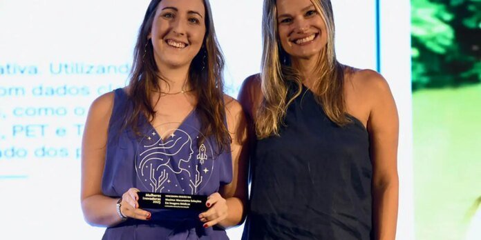 Two smiling women stand on stage, one holding a glass award plaque.