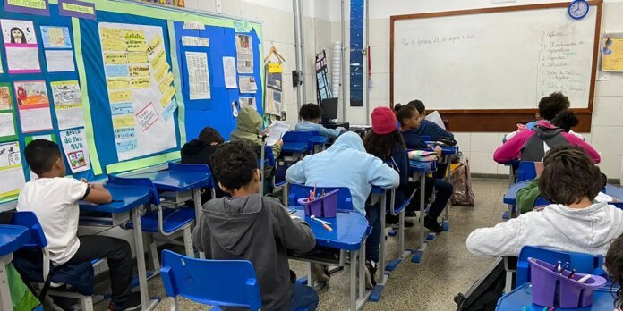 Students sit at blue desks in a classroom, working on worksheets with colorful bulletin boards on the walls behind them.