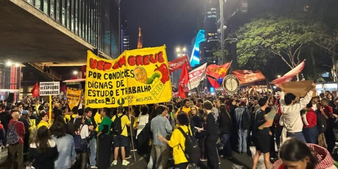 Night-time protest with a large yellow banner demanding dignified study and work conditions, crowd holding signs and flags across a city street.