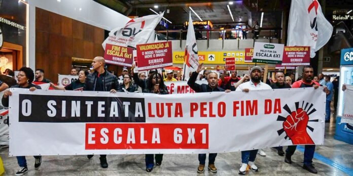 Grupo de manifestantes seguran uma faixa gigante branca com letras pretas e vermelhas pedindo o fim da escala 6x1 durante um protesto em um shopping.