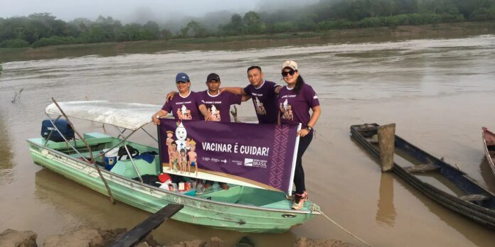 Five people in matching purple shirts stand on a green boat, holding a purple banner that says 'VACINAR É CUIDAR!' by a muddy river.