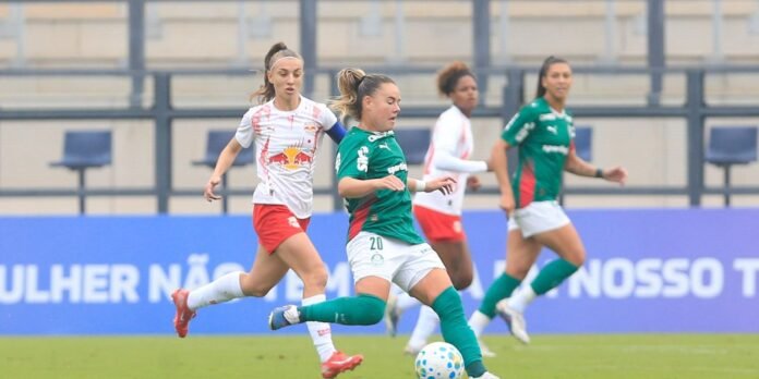 Women’s soccer match: a player in green kicks the ball while a white‑and‑red opponent pursues nearby on a grassy field with a blue sideline barrier behind them.