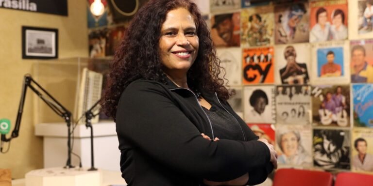 Smiling woman with curly hair and a black jacket stands with arms crossed in a radio studio, wall of vinyl records behind her.