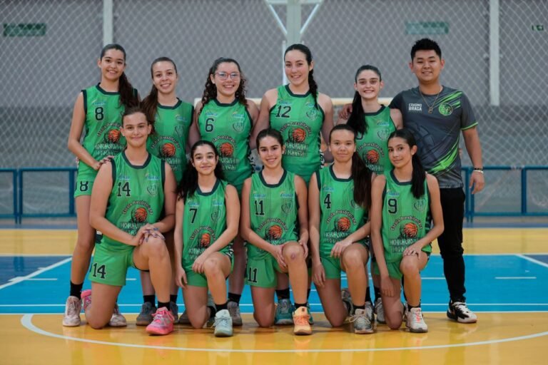Group photo of a girls' basketball team in green jerseys posing on an indoor court with their coach nearby in a gray shirt.