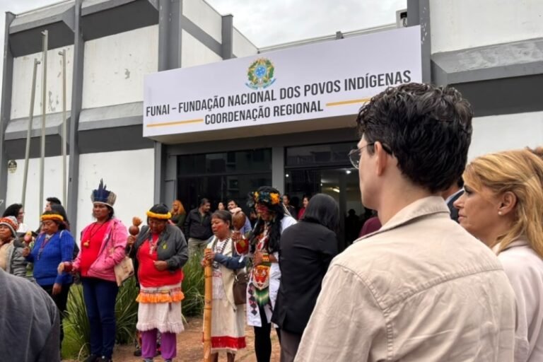Group of Indigenous people in colorful traditional attire stand outside a building with a FUNAI regional sign.