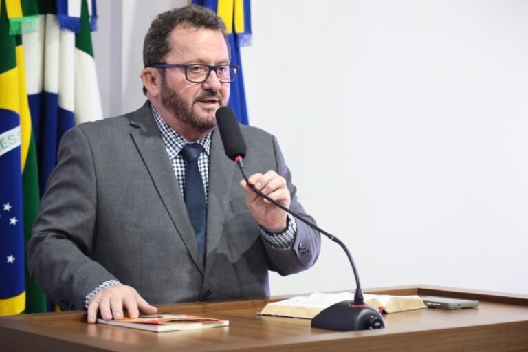 Man in a gray suit and glasses speaks into a microphone at a wooden podium, with flags in the background.