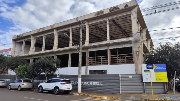Unfinished concrete building with open bays and exposed framework behind a metal fence on a city street. Cars are parked in front.