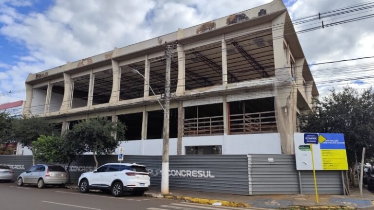 Unfinished concrete building with open bays and exposed framework behind a metal fence on a city street. Cars are parked in front.