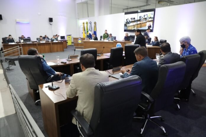 Formal meeting in a large conference room with delegates seated around a curved table, flags and screens in the background.