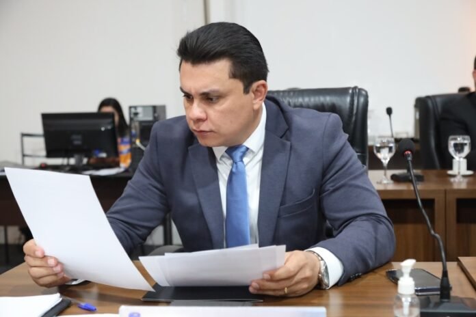 Man in a navy suit and blue tie reviews documents at a conference table during a meeting as others work in the background.