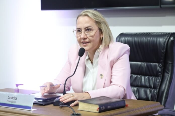 Woman in a pale pink blazer speaks into a microphone at a panel, seated at a wooden desk.