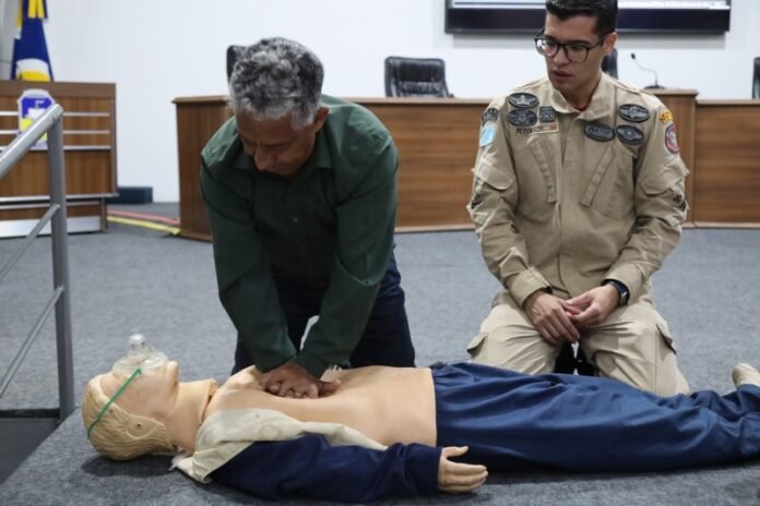 Man in a green shirt performs chest compressions on a CPR manikin while another person in uniform observes during a training session in a classroom-like setting