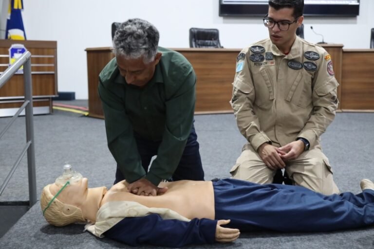 Man in a green shirt performs chest compressions on a CPR manikin while another person in uniform observes during a training session in a classroom-like setting