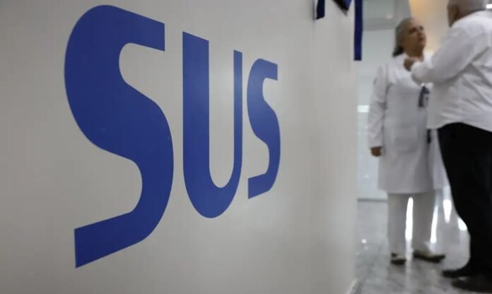 Large blue 'SUS' sign on a white wall, with two healthcare professionals in white coats talking in a hospital hallway behind it.
