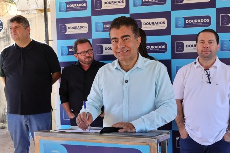 Man in a light blue shirt signs a document at a podium while two men stand behind him in front of a blue backdrop labeled 'Dourados Prefeitura'.