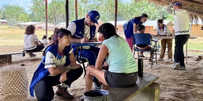 Volunteer in a blue vest speaks with a seated woman at an outdoor community clinic under a thatched shelter.