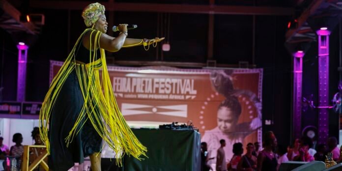 Singer performing on stage in a black outfit with bright yellow fringe shawls and a patterned headwrap, holding a microphone.