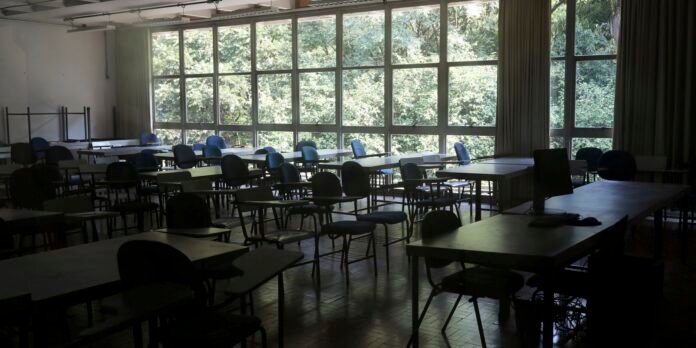 Empty classroom with rows of desks and chairs, large windows showing greenery outside; a computer monitor on a desk to the right.