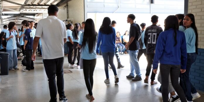 Group of students in a school hallway, many wearing light blue shirts, gathered and chatting near a bright entrance door.