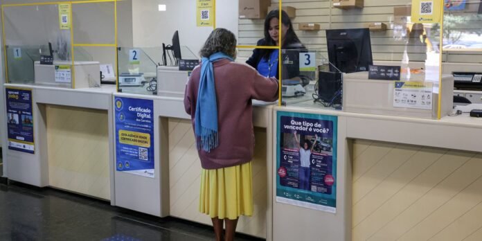 A customer at a service counter speaking with a clerk behind a protective glass partition in a bank or post office.