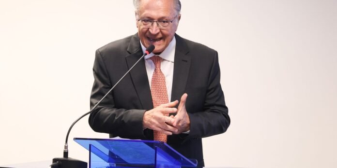 Senior man in a dark suit and red patterned tie speaks into a microphone at a blue lectern during a presentation.