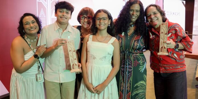 Six smiling people pose together, each holding tall carved trophy-style awards at an event backdrop.