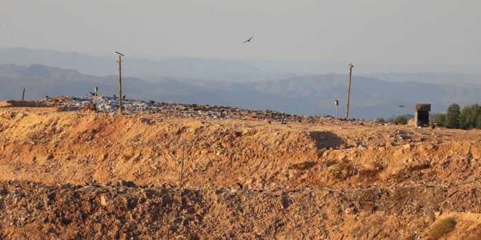 A barren landfill with mounded dirt and scattered piles of garbage under a hazy sky, with distant mountains in the background and a few birds in flight.