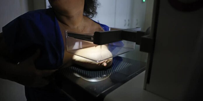 Healthcare worker in blue scrubs positions a woman's breast under a mammography machine for imaging.